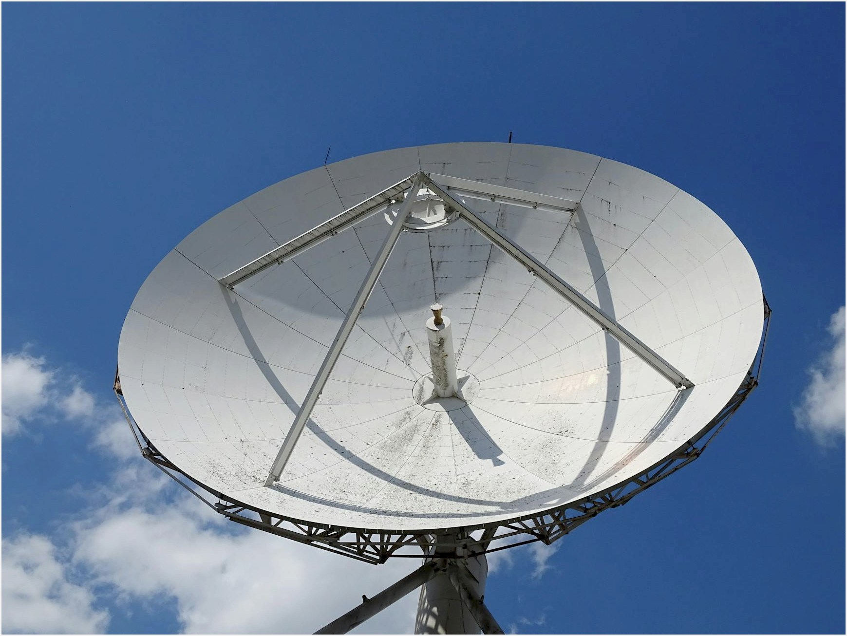 A low angle view of a large satellite dish against a clear blue sky with scattered clouds.