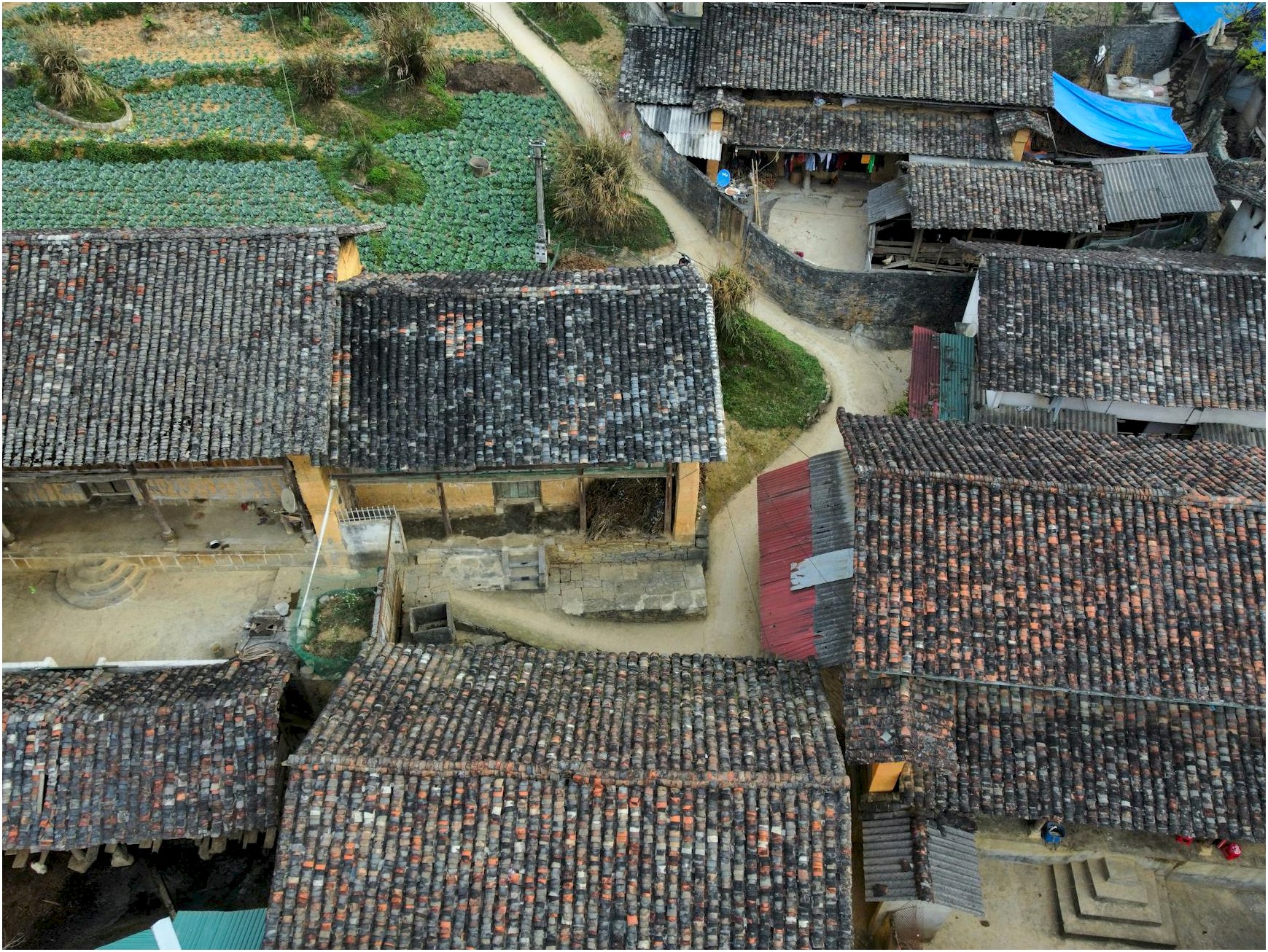 Aerial shot of a rustic village with traditional tiled roofs surrounded by farmland and pathways.