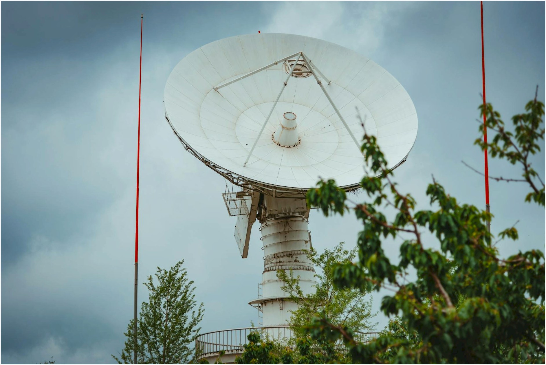 A large parabolic satellite dish with a cloudy sky background, surrounded by greenery.