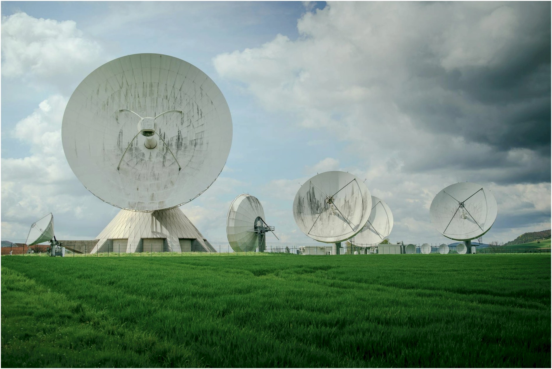 Array of large satellite dishes in a lush green field under a cloudy sky.